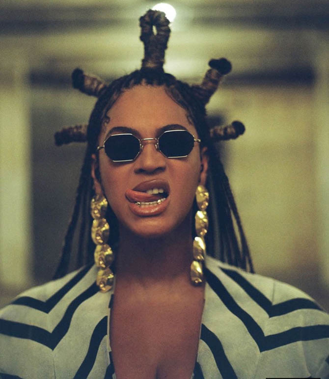 Beyonce wearing sunglasses and long gold earrings, licking her lips in Black Is King.