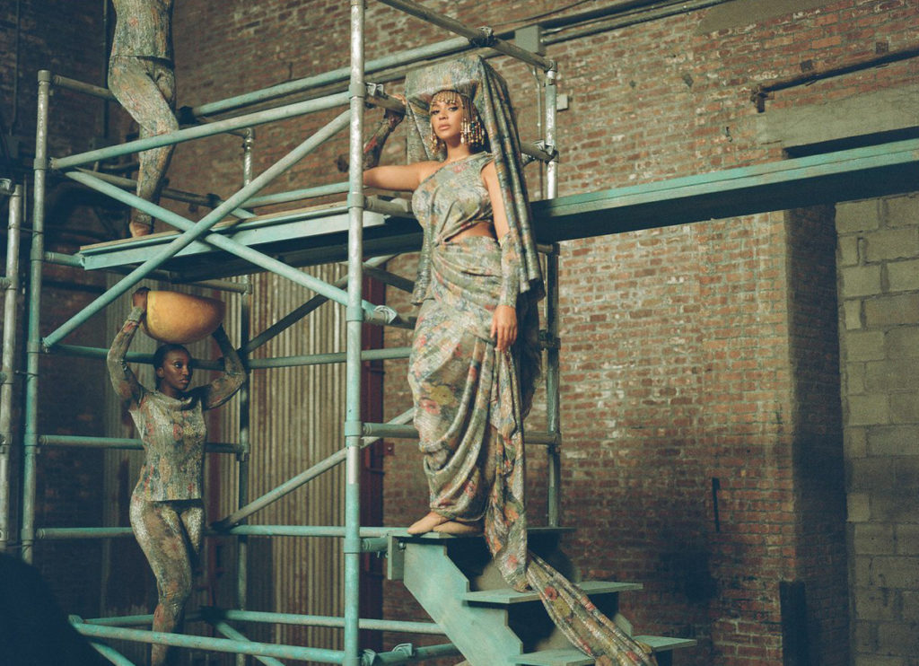 Beyonce wearing a drapey floral gown and square hat, standing on scaffolding, in Black Is King.